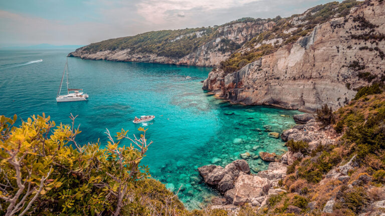 Eine malerische Küstenszene mit klarem, türkisfarbenem Wasser, Booten auf ruhigem Meer, felsigen Klippen und üppigem Grün. Der Himmel ist bedeckt und verleiht der Landschaft eine ruhige Atmosphäre. Die dramatischen Klippen bilden einen Kontrast zum lebhaften Wasser darunter und bilden eine ideale Kulisse für einen Segelurlaub an Bord einer Segelyacht.