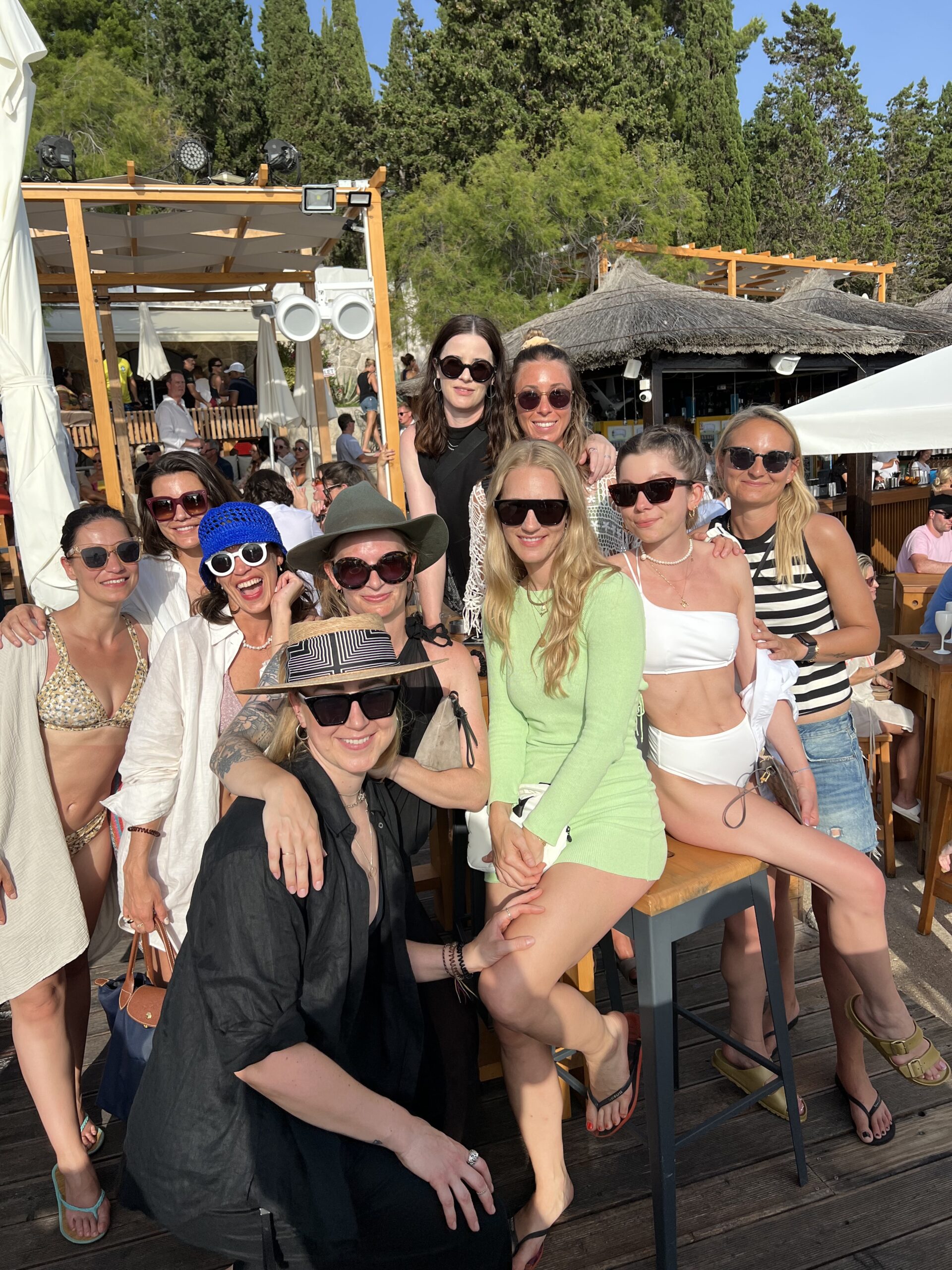 Eine Gruppe von Frauen genießt einen sonnigen Tag in einer Bar im Freien, lächelt und posiert für ein Foto, in Sommeroutfits gekleidet, mit Bäumen und einem Strohdach im Hintergrund während ihres Segeltörns.