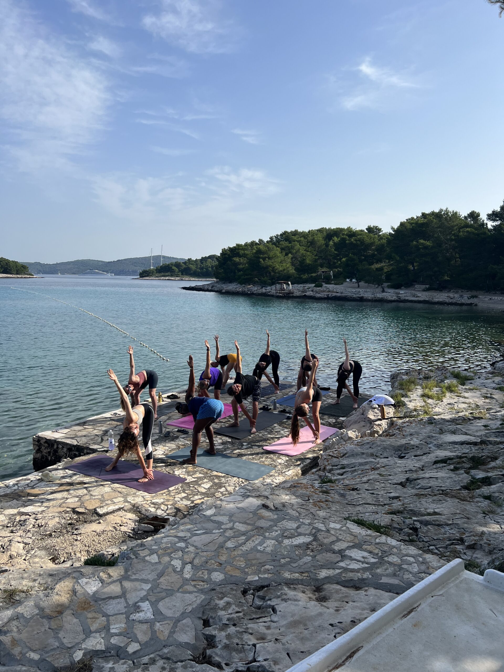 Eine Gruppe von Menschen praktiziert an einem sonnigen Tag Yoga auf einem Betonpier, umgeben von ruhigem Wasser und üppigem Grün, mit einer Segelyacht im Hintergrund.
