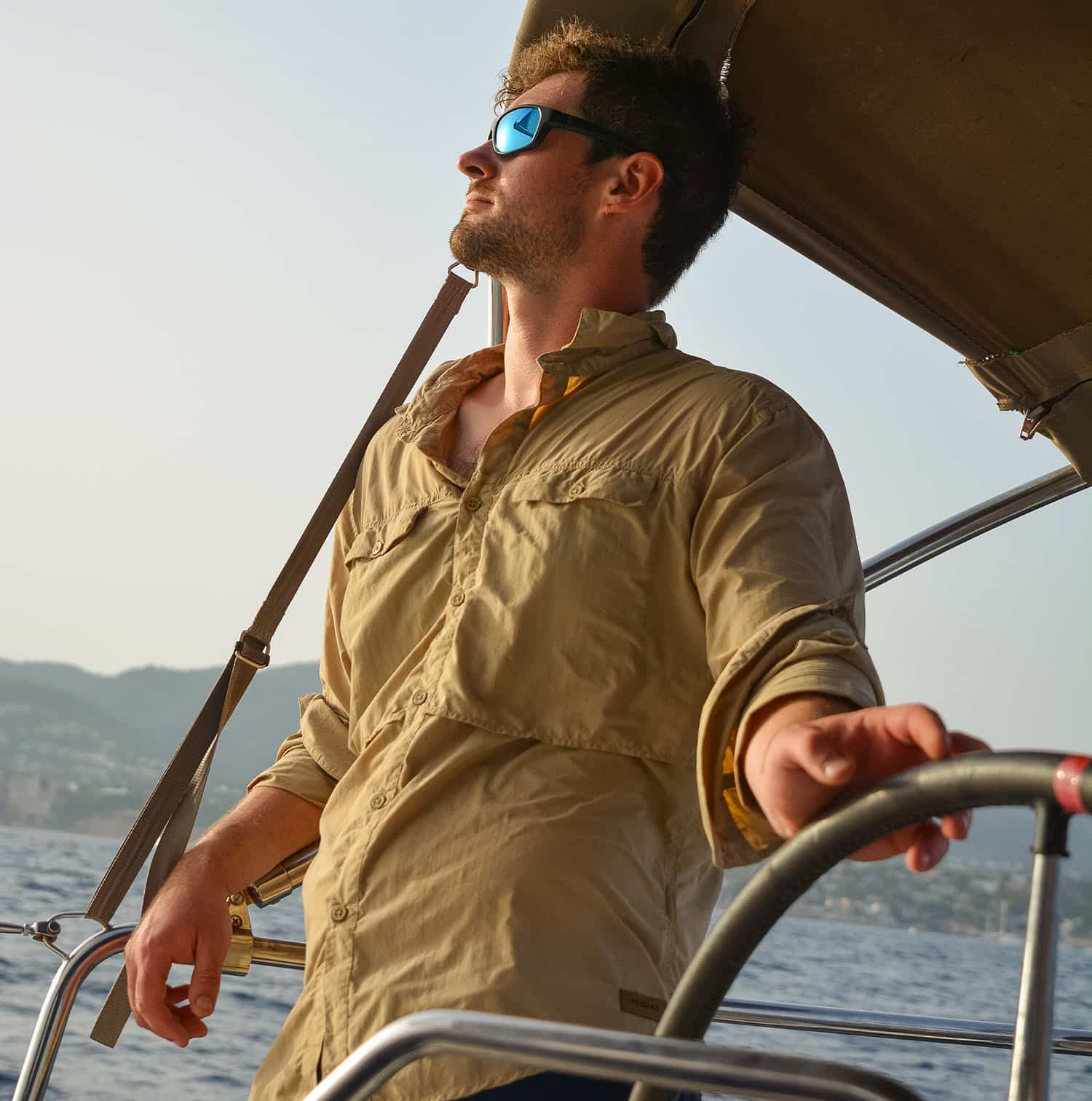 Ein Mann mit Sonnenbrille und beigem Hemd steuert mit konzentriertem Gesichtsausdruck einen Katamaran während des Sonnenuntergangs, im Hintergrund sind ruhiges Wasser und Hügel zu sehen.