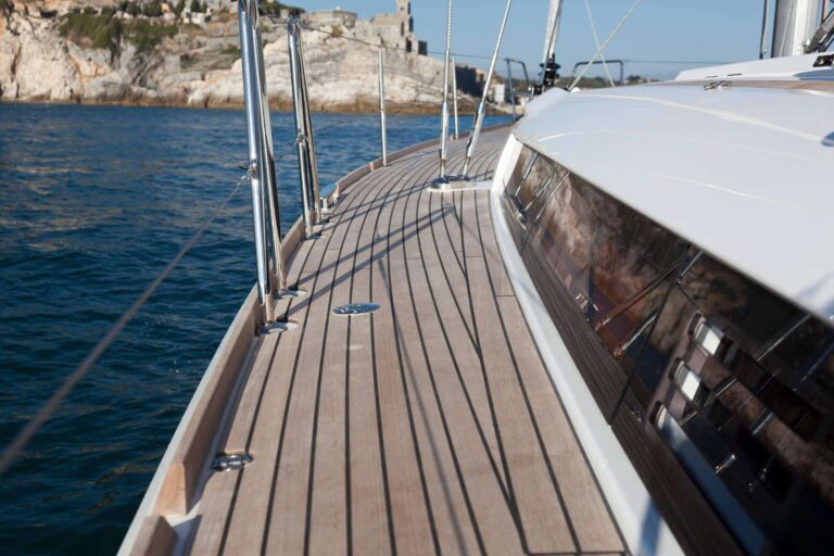 Blick vom Deck einer Segelyacht. Zu sehen sind Holzböden und Metallgeländer, im Hintergrund eine felsige Küste und klares blaues Wasser.