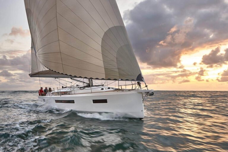 Ein weißes Segelboot mit großem Segel gleitet bei Sonnenuntergang über den Ozean. Der Himmel ist mit weichen, verstreuten Wolken bedeckt, die einen warmen Schein über die Szene werfen. Auf dem Deck der Segelyacht sind mehrere Personen zu sehen. Das Wasser wirkt leicht unruhig und spiegelt die goldenen Farbtöne des Himmels wider.