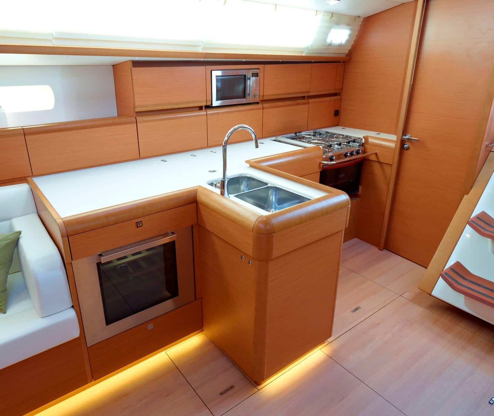 Bild einer modernen Bootsküche mit hellen Holzmöbeln, perfekt für eine Segelyacht. Sie verfügt über einen eingebauten Ofen, eine Mikrowelle, eine Doppelspüle und einen Gasherd. Der Raum ist gut beleuchtet und hat eine weiße Arbeitsplatte und einen gepolsterten Sitzbereich auf der linken Seite. Auf der rechten Seite sind Treppen zu sehen, die tiefer in die Segelyacht führen.