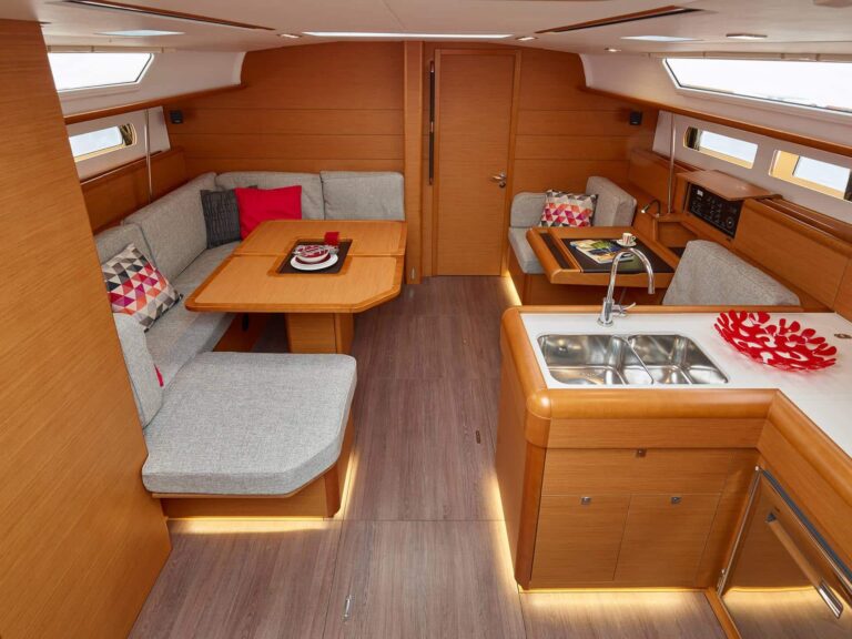 Ein modernes Segelyacht-Interieur mit Holzvertäfelung, einer gemütlichen Sitzecke mit grauen Polstern und bunten Kissen, einem Esstisch mit rotem Geschirr und einer kompakten Küche mit Spüle und rotem Dekoteller. Der Raum ist gut beleuchtet und stilvoll für eine unvergessliche Segelreise gestaltet.