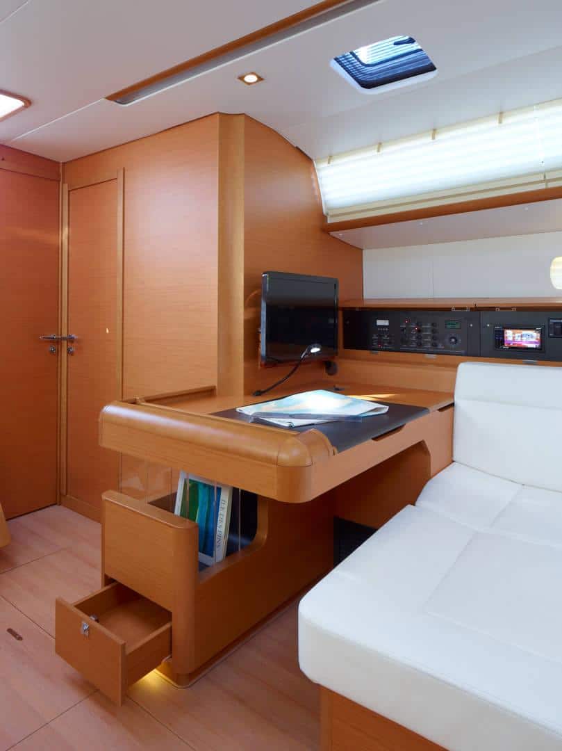 Eine moderne Bootskabine mit gepflegter Innenausstattung, perfekt für einen Segeltörn, mit einem Holzschreibtisch mit offener Schublade, einem weißen Sofa und Bedienfeldern an der Wand, die durch natürliches Licht von oben beleuchtet werden.