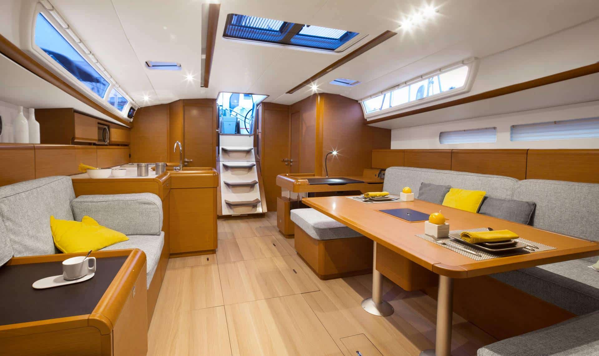 Innenansicht einer modernen Segelyacht mit gemütlichem Essbereich mit Holztisch und grauen Sofas, Küche mit Holzschränken und sonnendurchfluteten Oberlichtern.