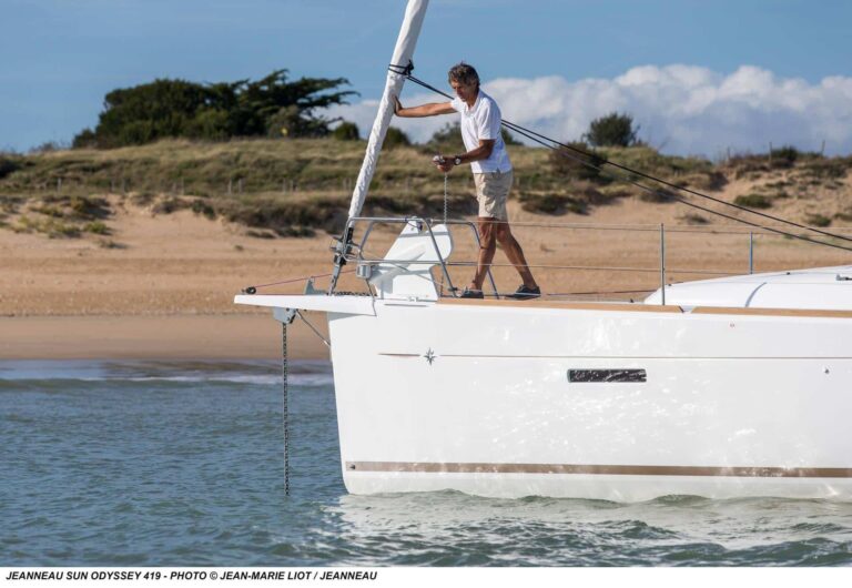 Eine Person in weißem Hemd und Shorts lichtet den Anker am Bug einer weißen Segelyacht Jeanneau Sun Odyssey 419. Das Boot liegt im seichten Wasser in der Nähe eines Sandstrands mit grasbewachsenen Dünen im Hintergrund und sorgt für einen idyllischen Segelurlaub unter einem klaren, sonnigen Himmel.
