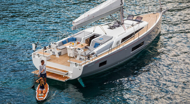 Ein Mann hilft einer Frau vom Achterdeck einer luxuriösen, modernen Segelyacht mit schnittigem Design auf ein aufblasbares Paddleboard. Das Boot liegt an einem sonnigen Tag in einem ruhigen, klaren blauen Meer vor Anker – perfekt für einen unvergesslichen Segelurlaub.