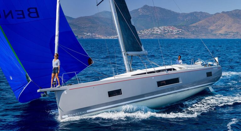 Ein schnittiges Segelboot mit weißem Rumpf und blauen und grauen Segeln gleitet durch das Wasser und begibt sich mit zwei Insassen an Bord auf einen friedlichen Segeltörn. Eine Person steht am Bug, während die andere vom Cockpit aus navigiert. Im Hintergrund sind Berge und eine Küstenstadt unter einem klaren Himmel zu sehen.