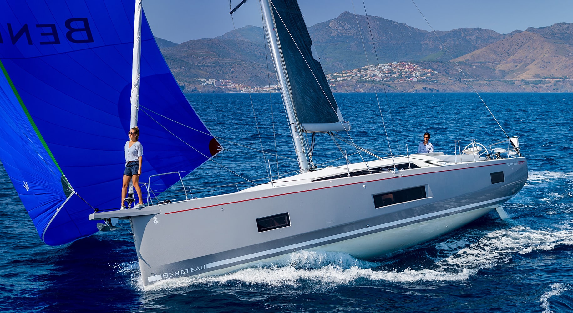 Eine schnittige Beneteau Segelyacht mit weißen und blauen Segeln gleitet durch das klare blaue Wasser. Zwei Personen sind an Bord, eine steht am Bug und die andere am Steuer. Im Hintergrund sind Berge und eine Küstenlinie zu sehen, die eine unvergessliche Segelreise versprechen.