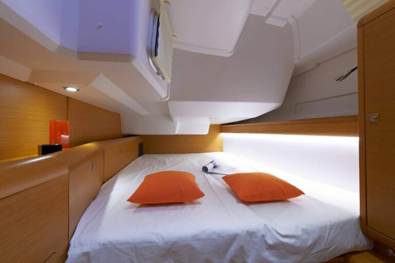Eine gemütliche, kompakte Kabine auf einem Katamaran mit einem Doppelbett mit weißer Bettwäsche und orangefarbenen Kissen, Holzvertäfelung und sanfter, stimmungsvoller Beleuchtung.