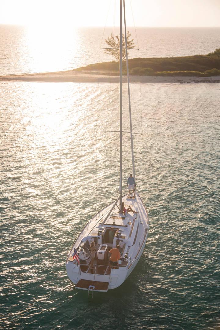 Eine Segelyacht mit einer Person an Deck bei Sonnenuntergang, die nahe einer bewaldeten Küste schwimmt, in das goldene Licht der untergehenden Sonne getaucht, während das Meer um sie herum schimmert.