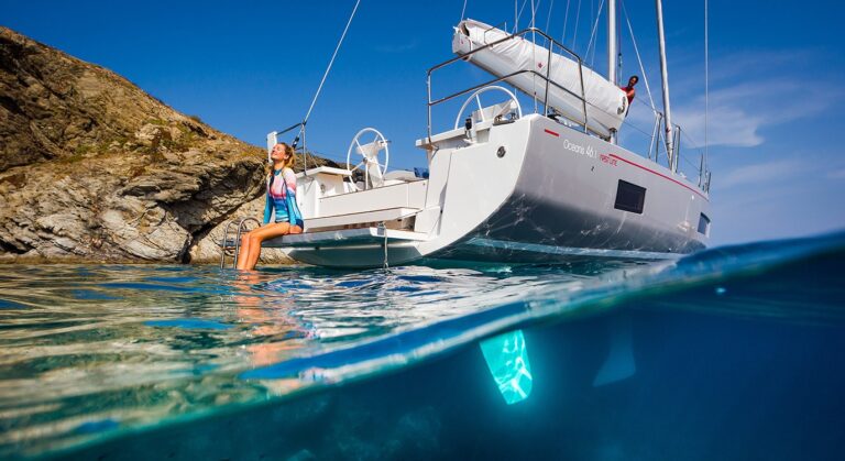 Eine Frau steht auf dem Heck eines Katamarans, der in der Nähe von Felsklippen vor Anker liegt. Im Vordergrund ist klares blaues Wasser zu sehen. Die Hälfte des Bildes zeigt eine Unterwasseransicht.