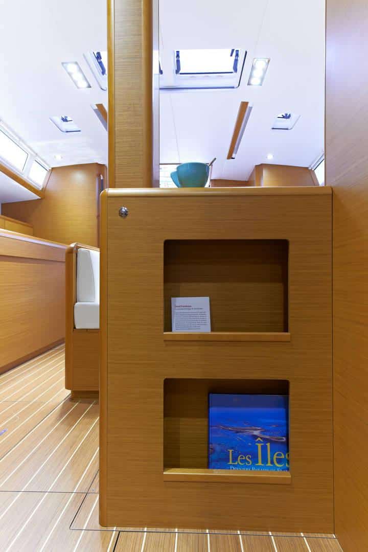 Innenansicht einer Segelyacht mit einem Holzschrank mit Regalen, die Bücher und eine dekorative Schale enthalten, unter einer gut beleuchteten Decke mit Holzpaneelen und einer Spiegelung auf einem polierten Boden.