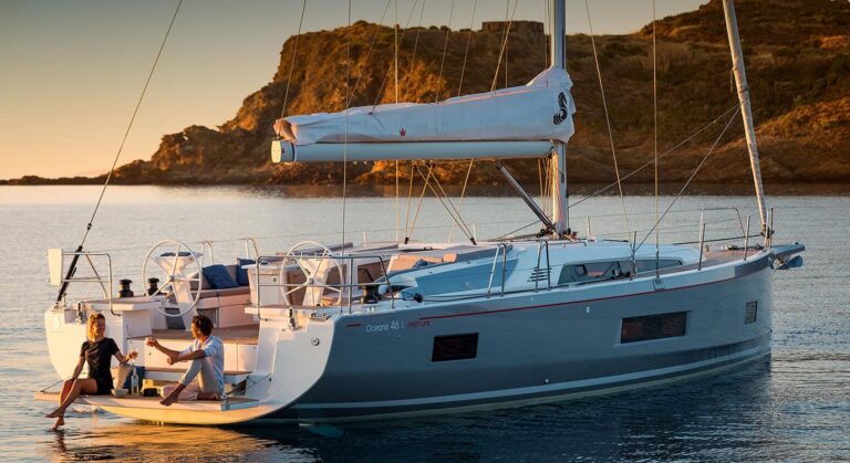 Eine Segelyacht liegt bei Sonnenuntergang in der Nähe eines Hügels vor Anker. Zwei Personen liegen hinten am Boot. Die untergehende Sonne wirft warmes Licht auf das ruhige Wasser und die raue Landschaft.