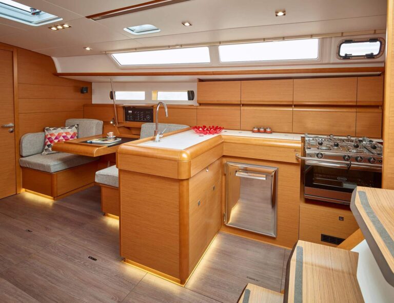 Eine moderne Segelyacht-Küche und ein Essbereich mit Holzschränken, einem Edelstahlherd und -ofen, einer Arbeitsplatte mit Spüle, einem kleinen Kühlschrank und einer gepolsterten Essecke mit Tisch, beleuchtet mit stilvollen Einbauleuchten. Perfekt für alle, die Mitsegeln-Abenteuer genießen.