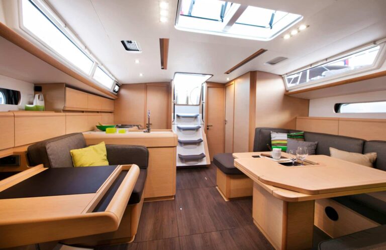 Innenansicht einer modernen Segelyachtkabine mit geräumigem Design, einem hölzernen Esstisch, gepolsterten Bänken, einer kompakten Küche und offenen Luken für natürliches Licht.