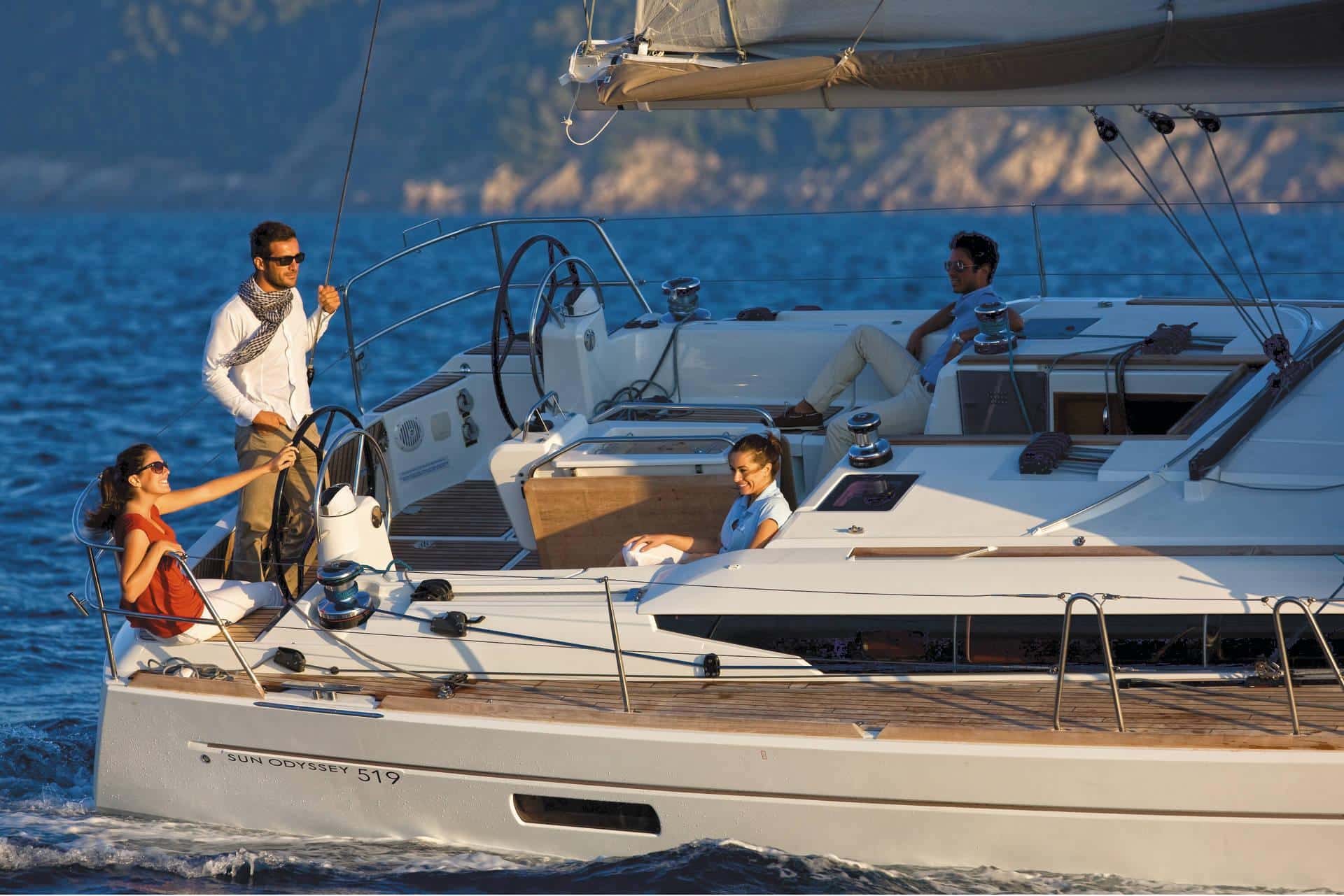 Eine Gruppe von Menschen genießt einen sonnigen Tag auf einem Segelboot namens „Sun Odyssey 519“ auf dem Wasser. Zwei Frauen sitzen auf dem Deck, während ein Mann am Steuerrad steht und eine andere Person sich am Heck des Bootes entspannt. Dieser perfekte Ausflug zeigt, warum Yachtcharter ein so begehrtes Erlebnis ist.