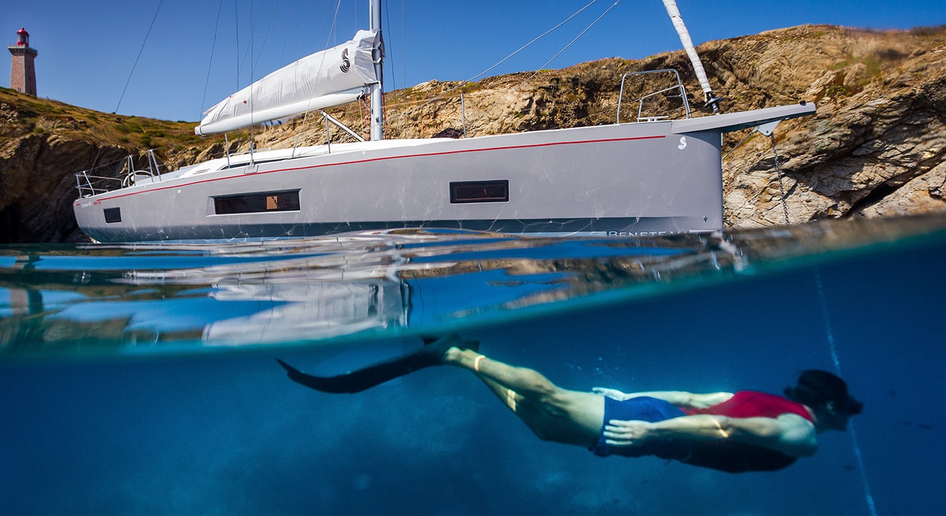 Eine Segelyacht ankert in der Nähe einer felsigen Küste über einem klaren, blauen Meer. Unter der Wasseroberfläche ist ein Mann in roten Shorts zu sehen, der schwimmt.