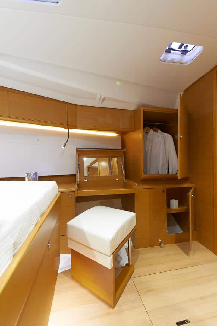 Innenansicht einer Yachtkabine mit warmer Holzvertäfelung, einem ordentlich gemachten Bett, einem offenen Kleiderschrank mit Kleidung, einem kleinen Schreibtisch mit Stuhl und sanfter Beleuchtung für einen gemütlichen Segelurlaub.