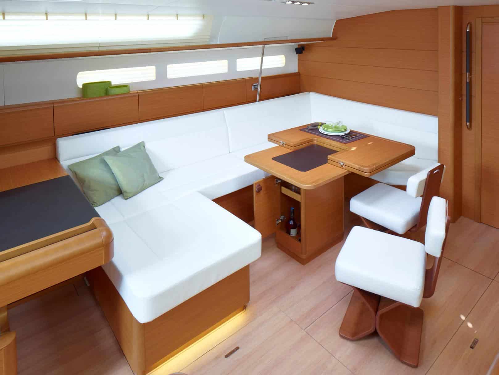Ein modernes Bootsinterieur mit Holzoptik und weißen Polstern, perfekt für ein Mitsegeln. Der L-förmige Sitzbereich ist mit grünen Kissen geschmückt. Ein kompakter Esstisch mit zwei Stühlen und ein kleiner Schrank sind ebenfalls vorhanden. Der Raum ist durch natürliches Licht aus den Fenstern gut beleuchtet, ideal zum Mitsegeln.