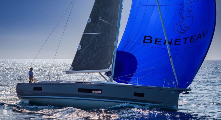 Ein Beneteau-Katamaran mit einem großen blauen Spinnaker segelt unter klarem Himmel über die ruhige See, während ein Mann am Bug steht und die Aussicht genießt.
