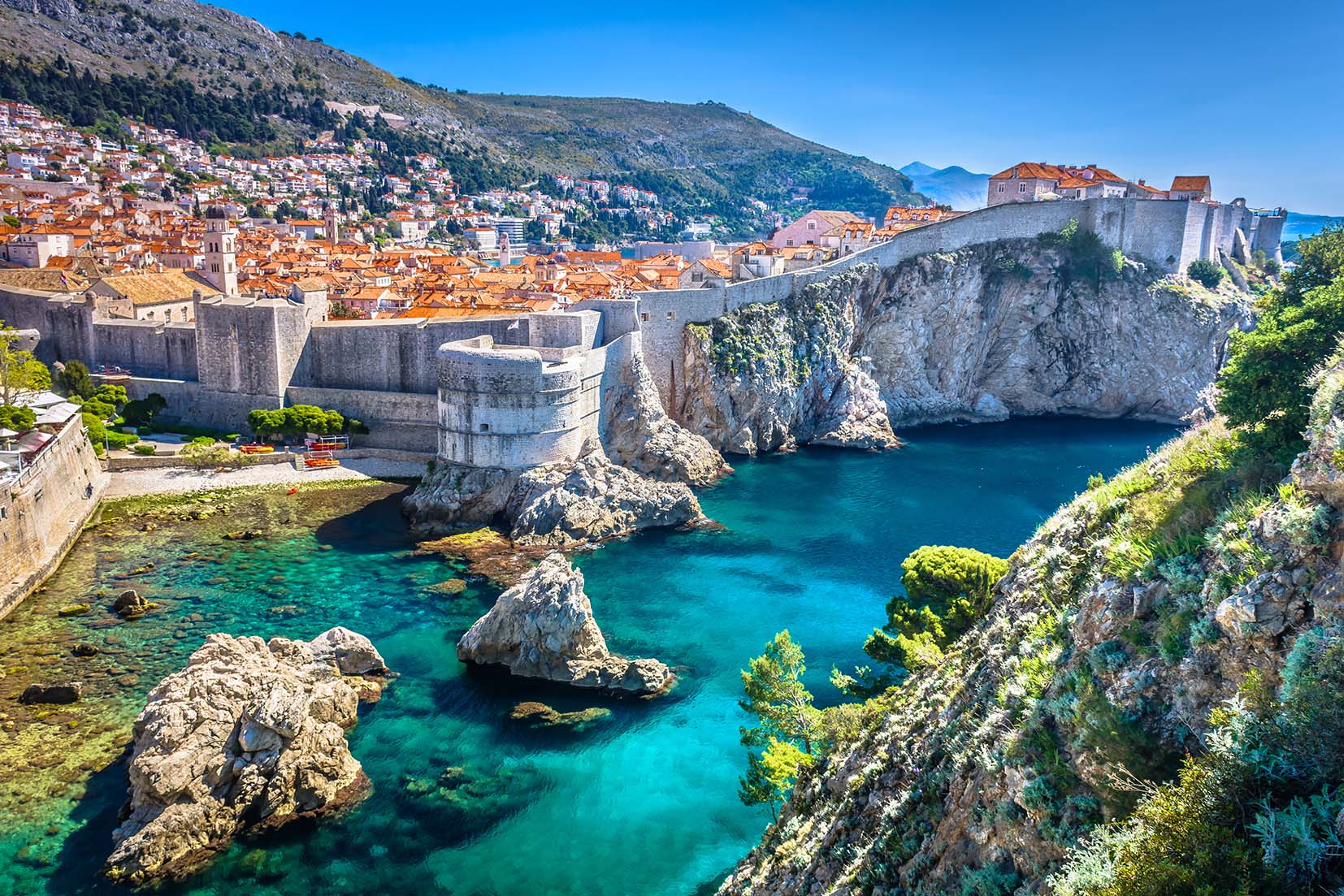 Ein Panoramablick auf Dubrovnik in Kroatien. Zu sehen sind die historischen Festungsmauern, das klare, türkisfarbene Wasser der Adria mit ihren vielen Segelyachten und die dicht gedrängten Gebäude mit Terrakotta-Dächern in der Altstadt.