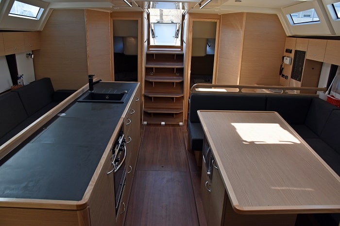 Innenansicht einer modernen Segelyachtkabine mit einer Küche mit langer Theke auf der linken Seite, einem Essbereich mit Tisch auf der rechten Seite und einer Holztreppe zum Oberdeck, alles umgeben von holzgetäfelten Wänden.