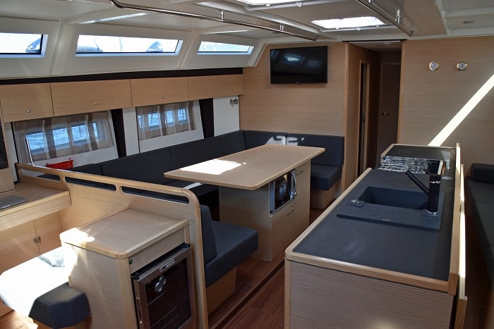 Innenansicht einer modernen Segelyacht mit einem ordentlich eingerichteten Essbereich mit einem Holztisch, umgebenden Sitzgelegenheiten, einer Küchenzeile mit Geräten und reichlich natürlichem Licht durch die Luken oben.