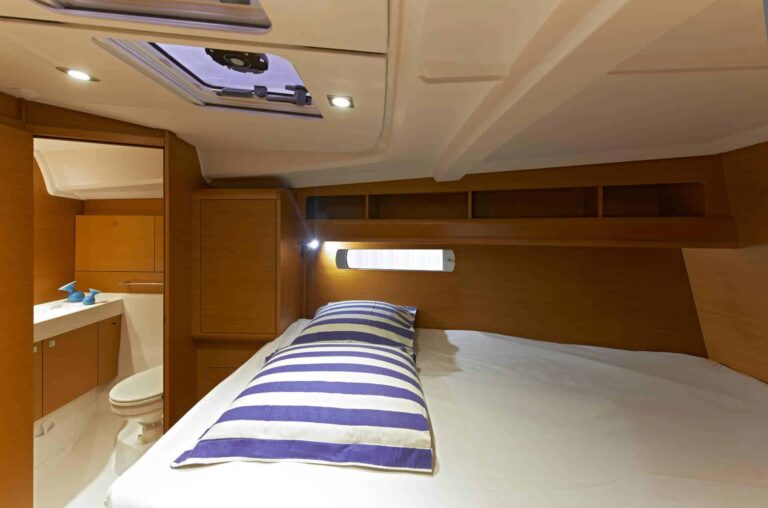 Das Bild zeigt das Innere einer Bootskabine mit einem gemütlichen Bett mit blau-weiß gestreiftem Kissen. Angrenzend befindet sich ein kleines Badezimmer mit Toilette und Waschbecken. Oben gibt es eine Luke und eingebaute Lichter, die den holzgetäfelten Raum erhellen – perfekt für Ihren nächsten Segelurlaub.