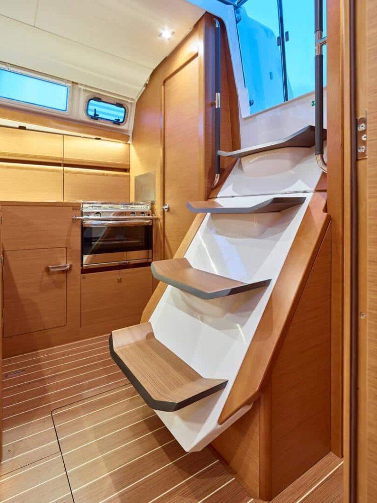 Innenansicht einer modernen Bootskabine mit Holzverkleidung. Das Bild zeigt eine schmale Treppe nach oben, daneben befindet sich ein kleiner Küchenbereich mit Herd, Schränken und Schubladen. Der Raum ist ideal für Ihren nächsten Segeltörn, ist mit Deckenbeleuchtung gut beleuchtet und strahlt sowohl Komfort als auch Stil aus.