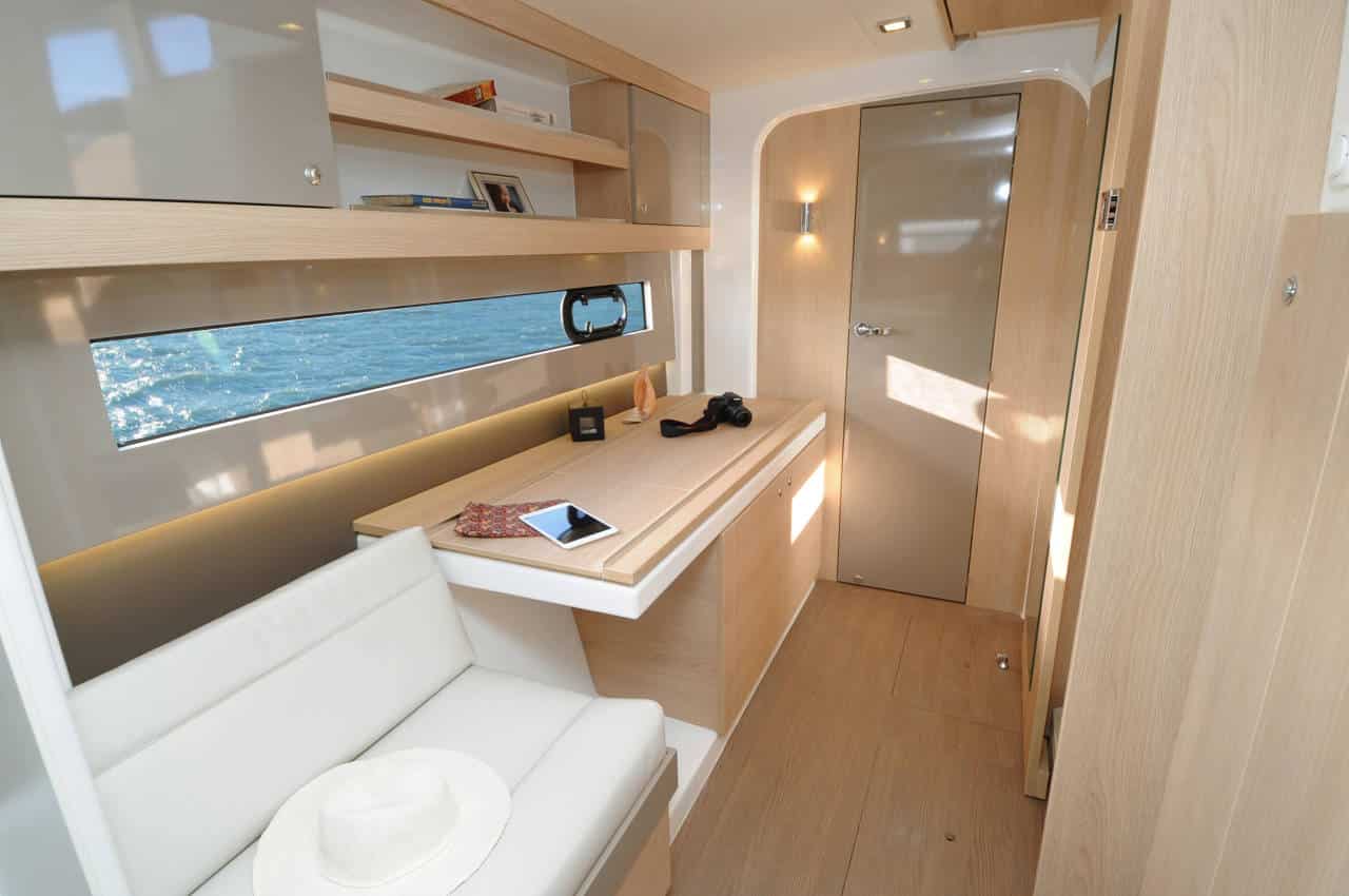 Innenansicht einer modernen Yachtkabine mit Holzverkleidung, einem kleinen weißen Sofa, einem Schreibtisch mit Tablet und Kamera, Bücherregalen und einer Tür, durch deren Fenster während des Segelurlaubs ein malerischer Meerblick sichtbar ist.