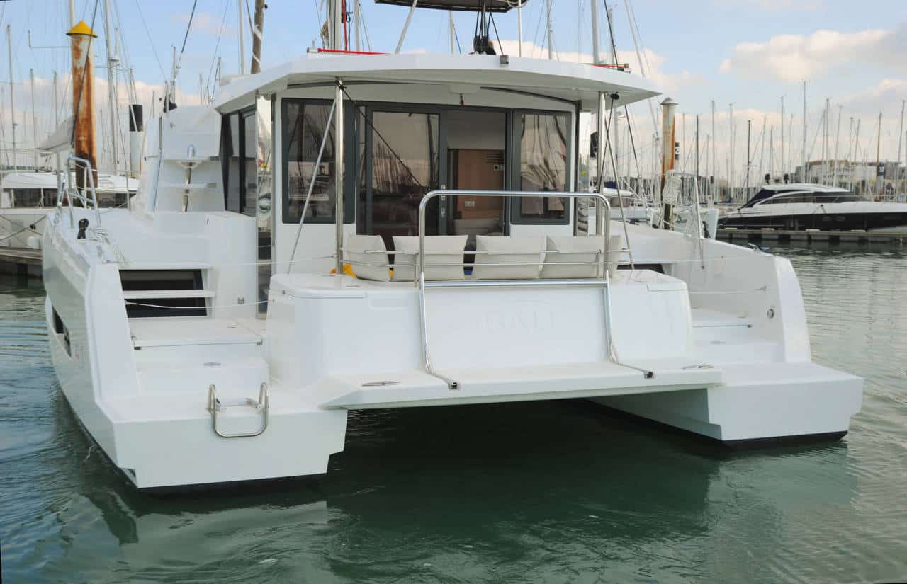 Eine moderne Segelyacht liegt in einem Yachthafen. Im Hintergrund sind unter einem bewölkten Himmel die ordentlich angeordneten Masten anderer Boote zu sehen.