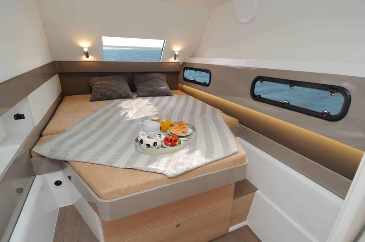Innenansicht einer Segelyachtkabine mit einem ordentlich gemachten Bett mit Kissen und einem Frühstückstablett mit Kaffee und Obst darauf, beleuchtet durch natürliches Licht aus Bullaugenfenstern.