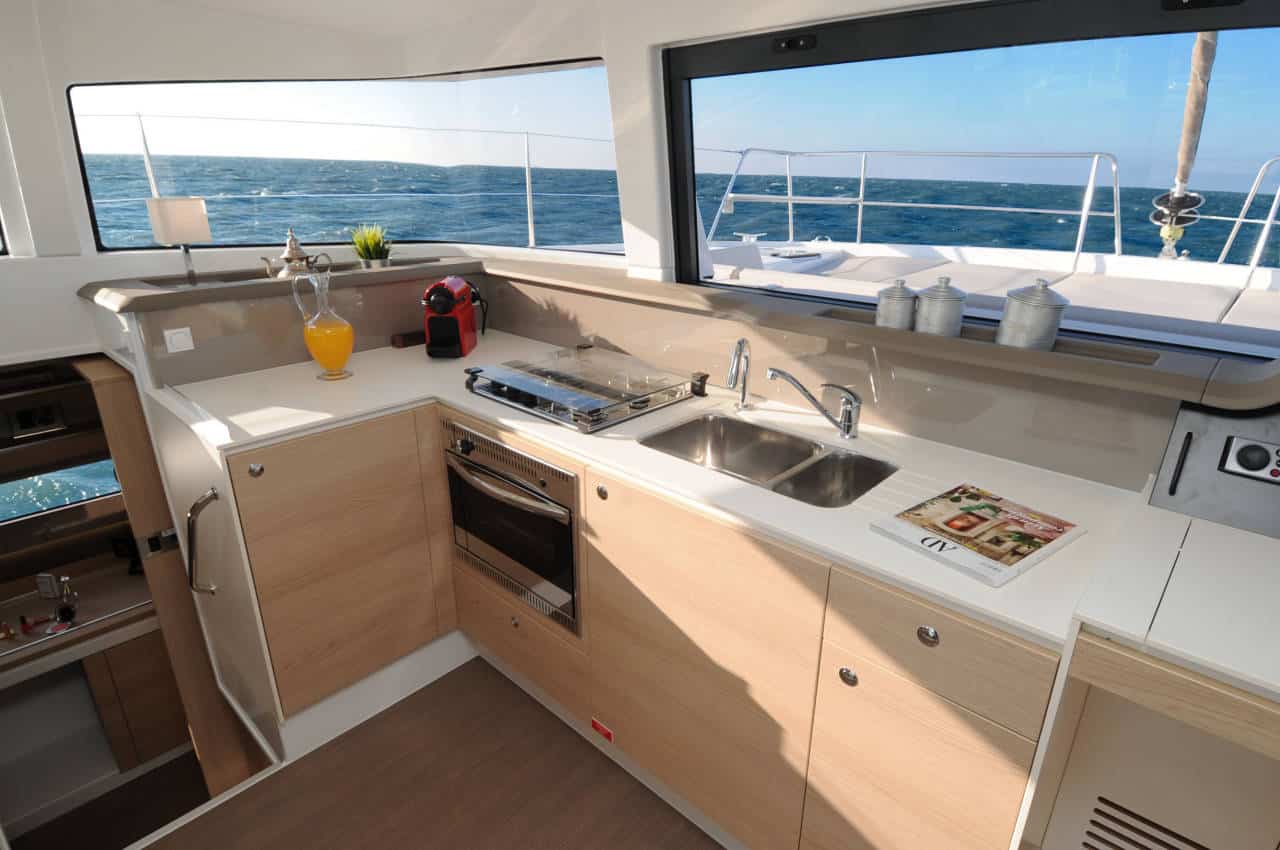 Eine moderne Segelyacht-Küche mit Holzschränken, Edelstahlspüle, Ofen und Arbeitsflächen. Helles Sonnenlicht strömt durch große Fenster herein und bietet Ausblick auf das Meer. Auf der Theke stehen Saftkrüge und eine Zeitschrift.