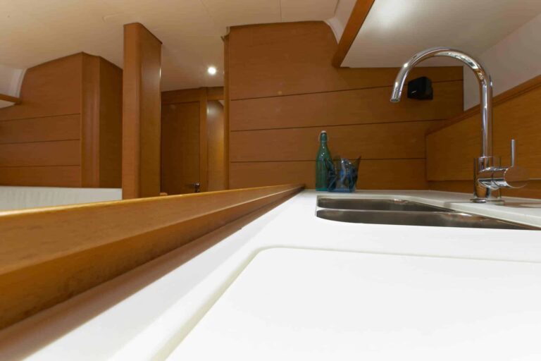 Modernes Kücheninterieur mit Holzschränken und einer sauberen Arbeitsplatte mit Edelstahlspüle und hohem Wasserhahn. Eine blaue Glasflasche verleiht einen dekorativen Touch, der an eine Segelreise erinnert.