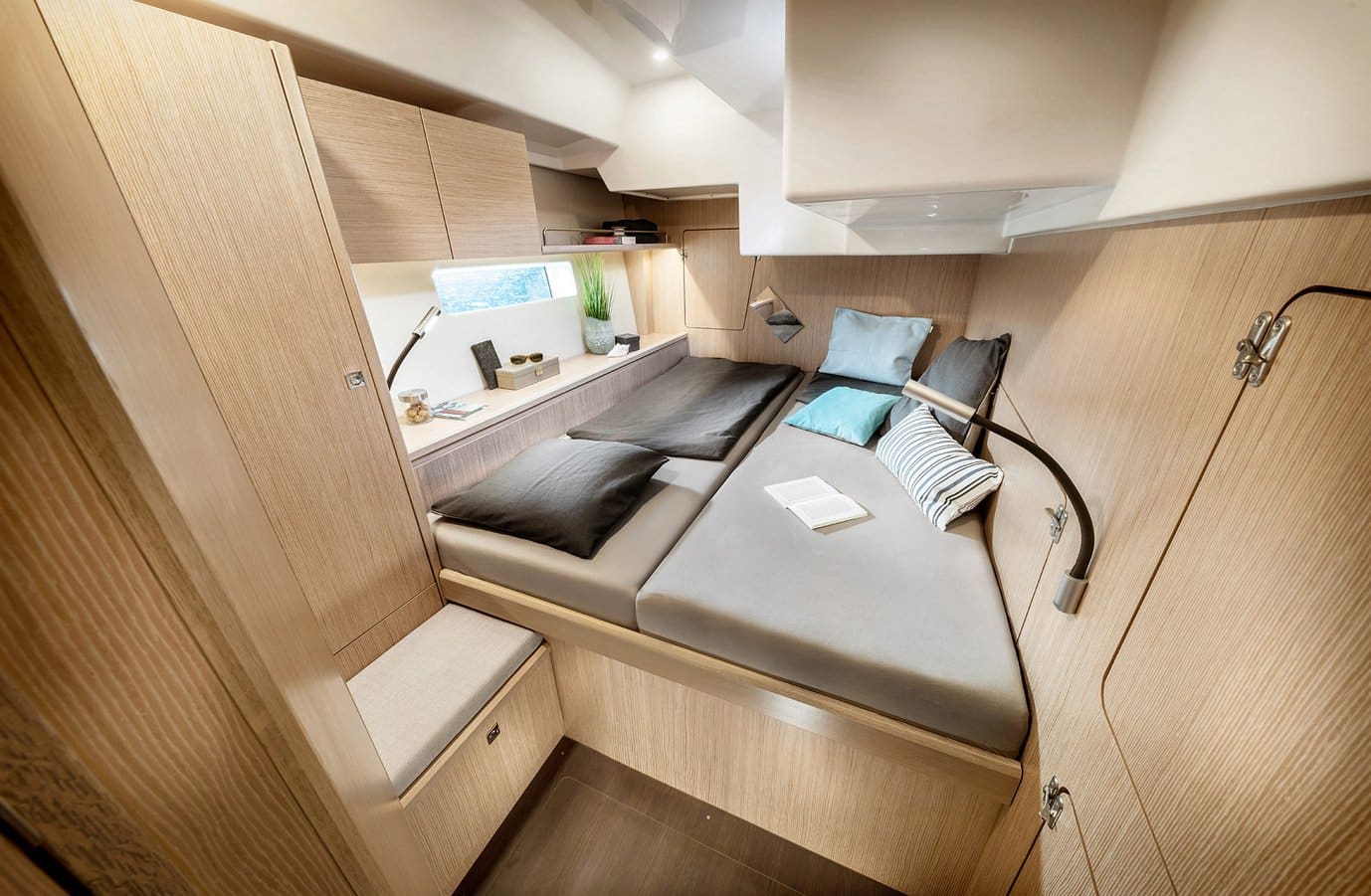 Modernes Schlafzimmer mit heller Holzvertäfelung, zwei Einzelbetten, jedes mit schwarzen und blauen Kissen, und einem Buch auf einem Bett. Eingebaute Schubladen und Schränke bieten Stauraum, während an der Wand angebrachte Leselampen für Beleuchtung sorgen. Ein Fenster erhellt den Raum und erinnert an eine gemütliche Segelurlaubshütte.