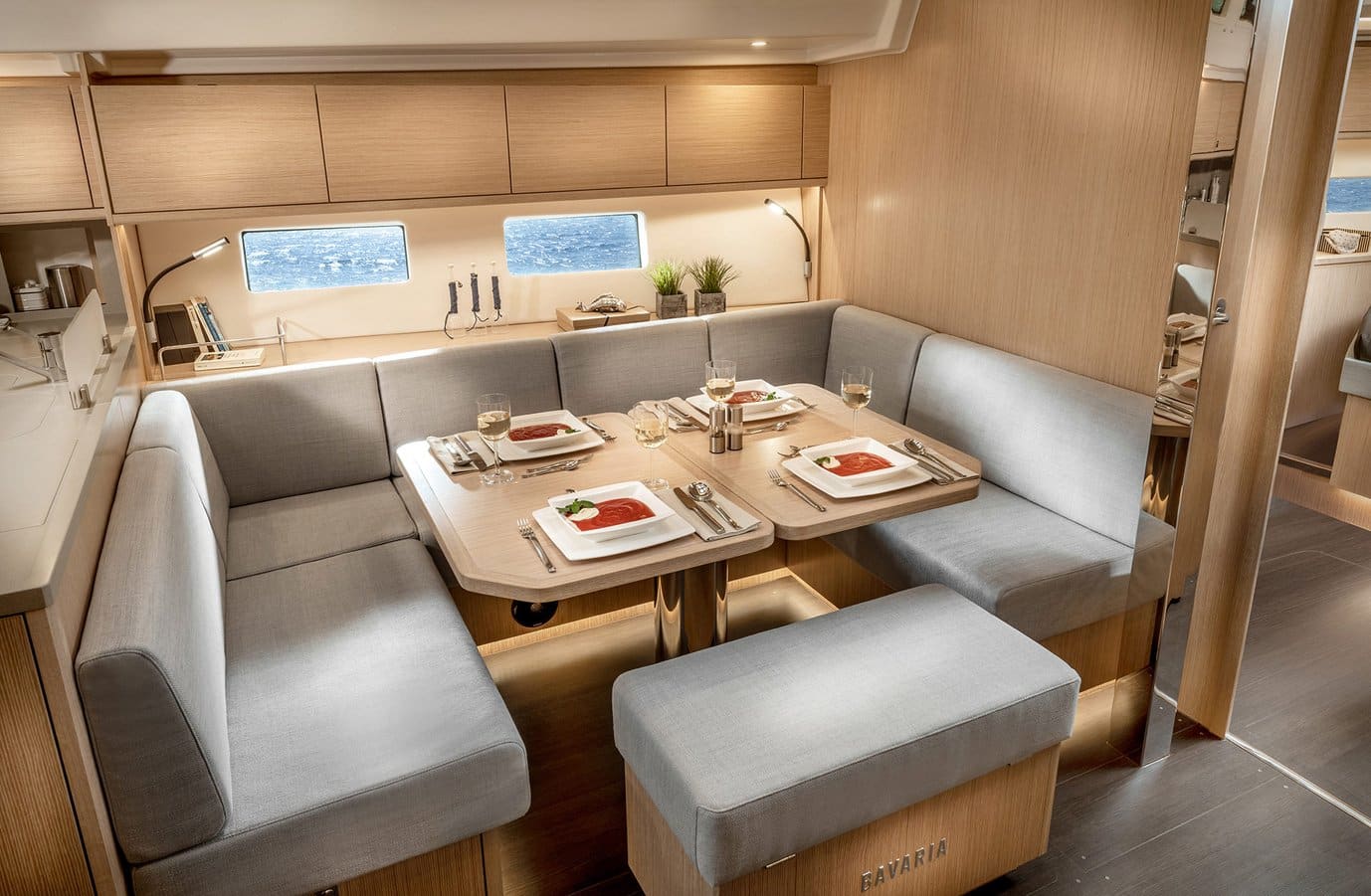 Innenansicht eines modernen Essbereichs in einem Katamaran mit einem U-förmigen grauen Sofa, einem Holztisch für zwei Personen, zusätzlichen Hockern und großen Fenstern mit Meerblick.