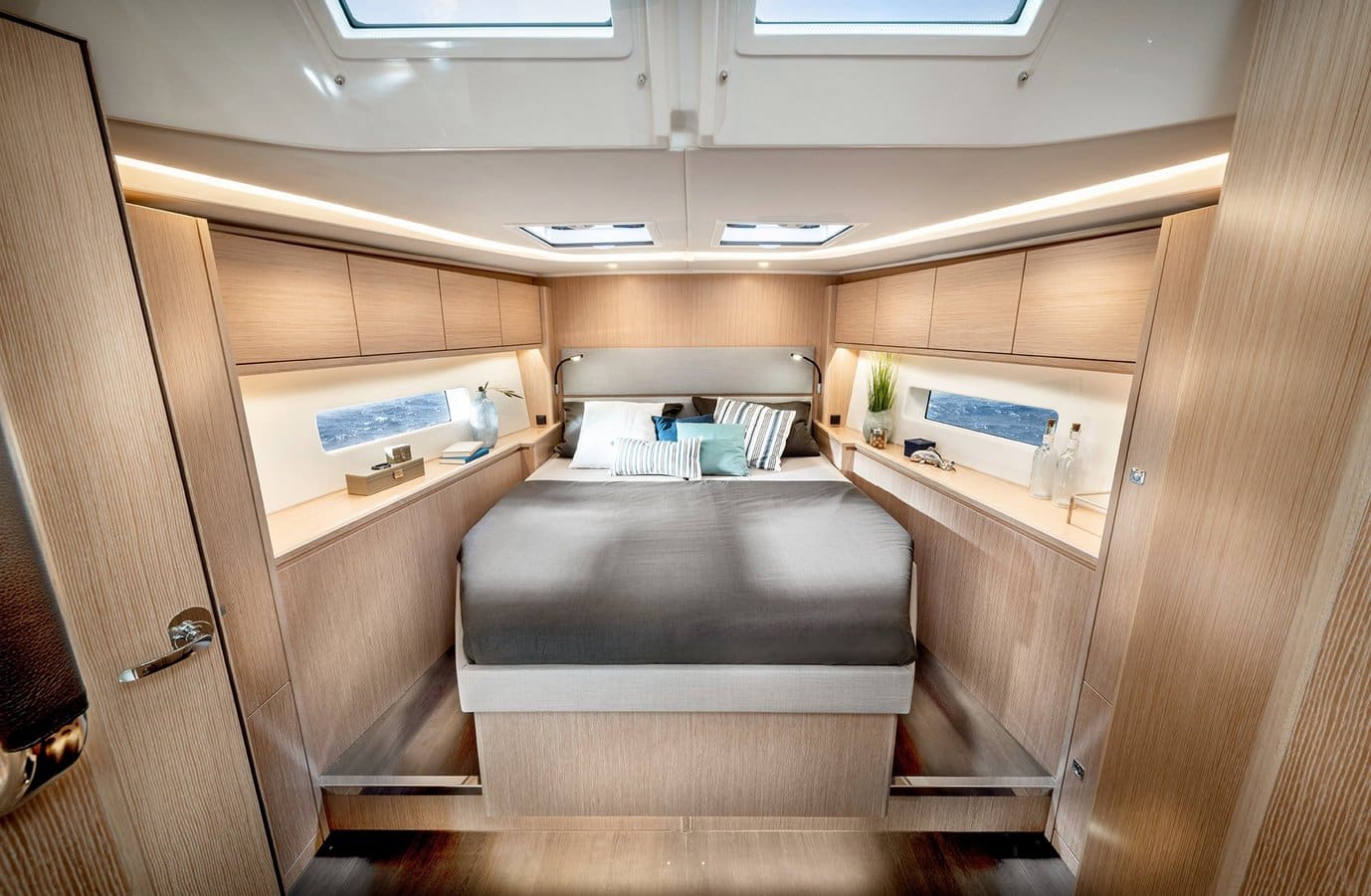 Innenansicht eines luxuriösen Schlafzimmers auf einer Segelyacht mit einem großen Bett in der Mitte, flankiert von Holzschränken und großen Fenstern auf beiden Seiten, die für ein helles und modernes Ambiente sorgen.