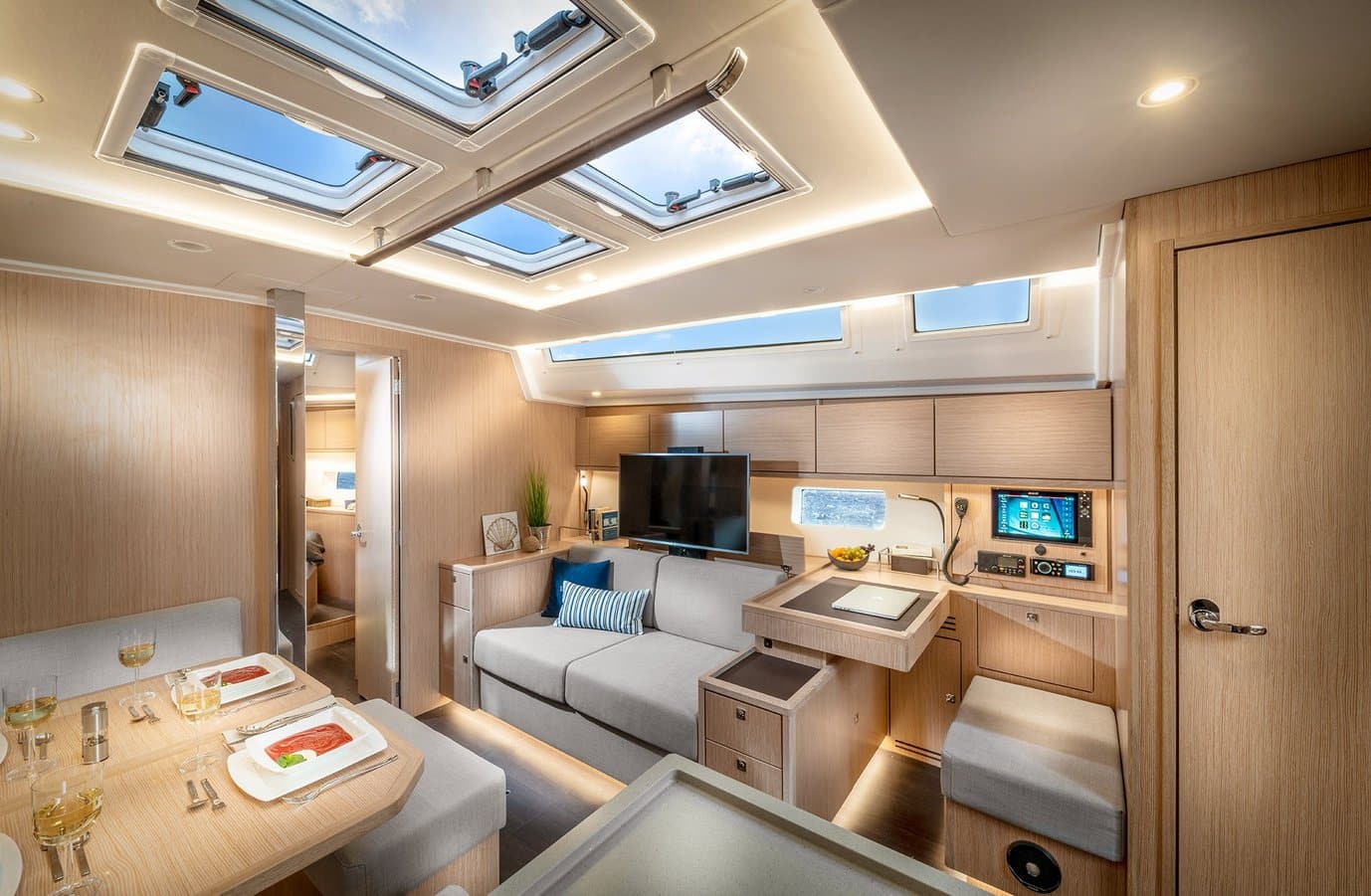 Innenansicht einer modernen Segelyachtkabine mit einem gemütlichen Sitzbereich mit Sofa und Esstisch, einer kompakten Küche mit Geräten und mehreren Oberlichtern für natürliches Licht.