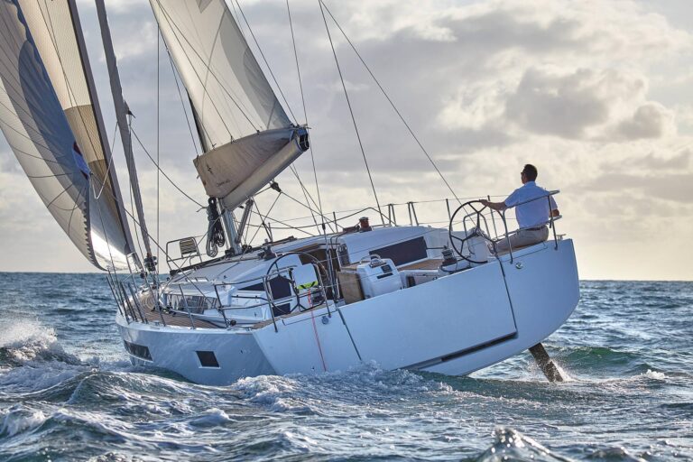 Ein Mann steuert eine große Segelyacht auf rauer See mit voll geöffneten Segeln unter einem bewölkten Himmel bei Sonnenuntergang. Das Boot ist aufgrund des starken Windes geneigt.