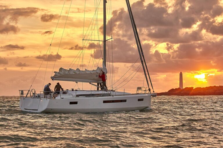 Eine Segelyacht mit eingerollten Segeln auf dem Meer bei Sonnenuntergang, mit zwei Personen an Deck und einem Leuchtturm im Hintergrund unter einem orangefarbenen Himmel.