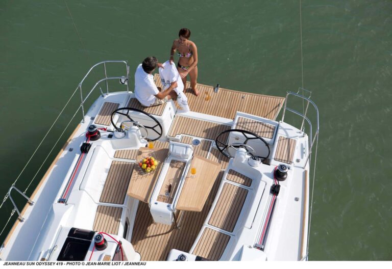 Zwei Personen genießen einen sonnigen Tag auf einer Luxus-Segelyacht. Der Mann steht am Steuer und die Frau sitzt daneben inmitten eines geräumigen Teakdecks. Wasser und Horizont sind sichtbar.