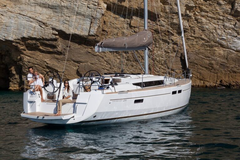 Eine moderne Segelyacht mit zwei Personen an Bord, eine davon am Steuer, in der Nähe einer Felsklippe unter klarem Himmel.
