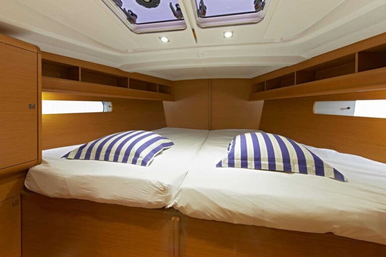 Innenansicht einer Katamaran-Kabine mit einem ordentlich gemachten Bett mit gestreiften Kissen, umgeben von Wänden und Schränken aus poliertem Holz, in das natürliches Licht durch die seitlichen Bullaugen fällt.