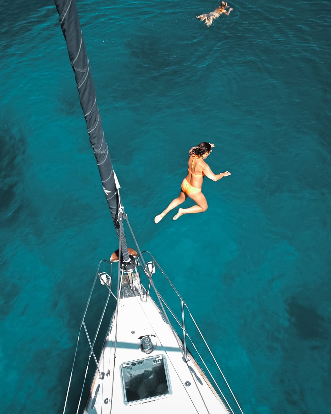 Eine Person springt von einem Segelboot ins klare blaue Wasser und genießt ihren Segelurlaub. Eine andere Person schwimmt im Hintergrund. Das von oben aufgenommene Foto zeigt den Mast des Bootes und einen Teil des Decks. Das ruhige und einladende Wasser verkörpert das ideale Segelreise-Erlebnis.
