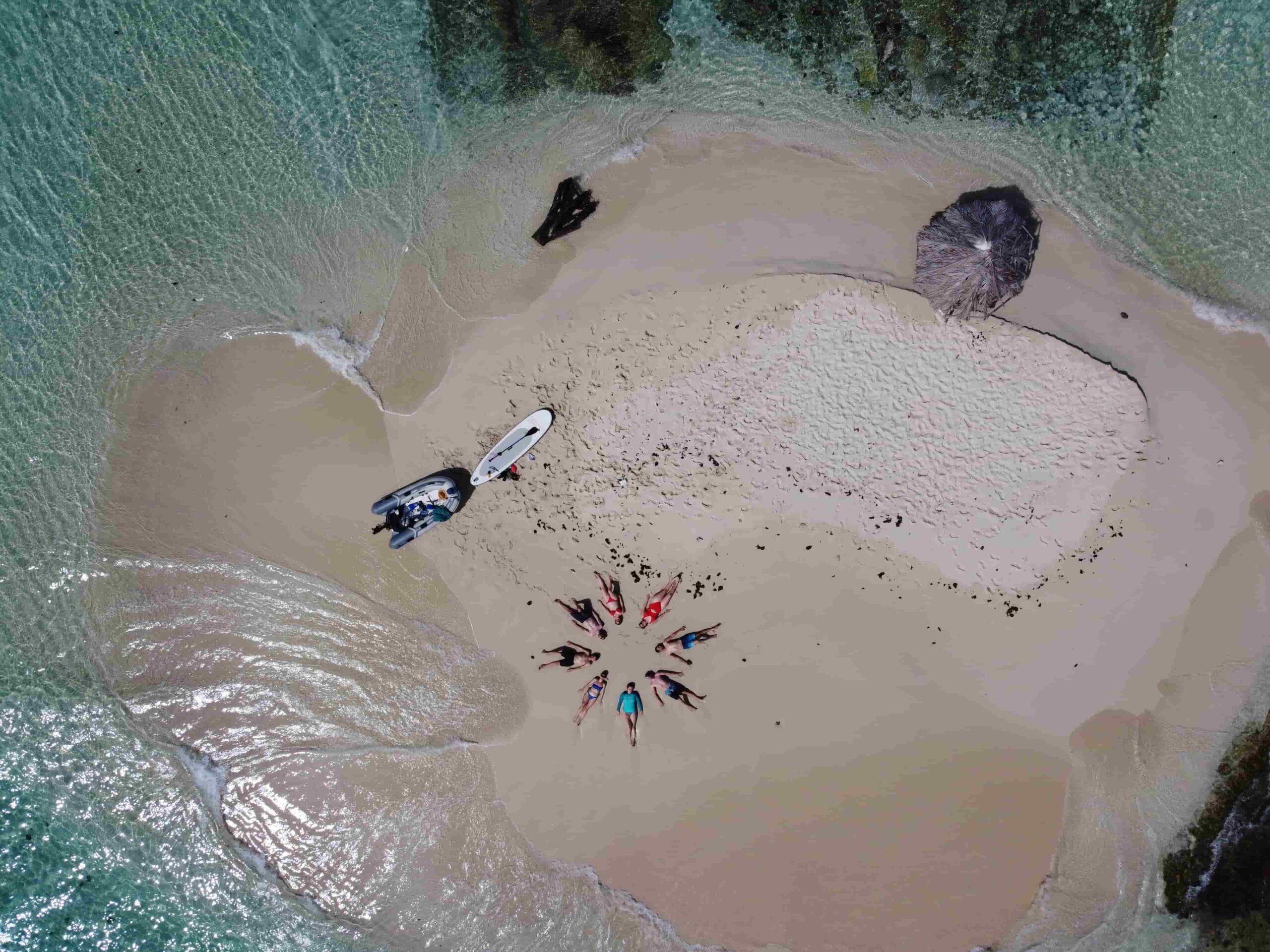 Luftaufnahme einer kleinen Sandinsel, umgeben von klarem, türkisfarbenem Wasser. Mehrere Menschen liegen kreisförmig im Sand, in der Nähe ein Kajak, ein Paddleboard und ein kleines Boot. Es ist eine idyllische Szene, perfekt für jede Kombinationsreise. Ein Sonnenschirm und Handtücher liegen verstreut am Strand.