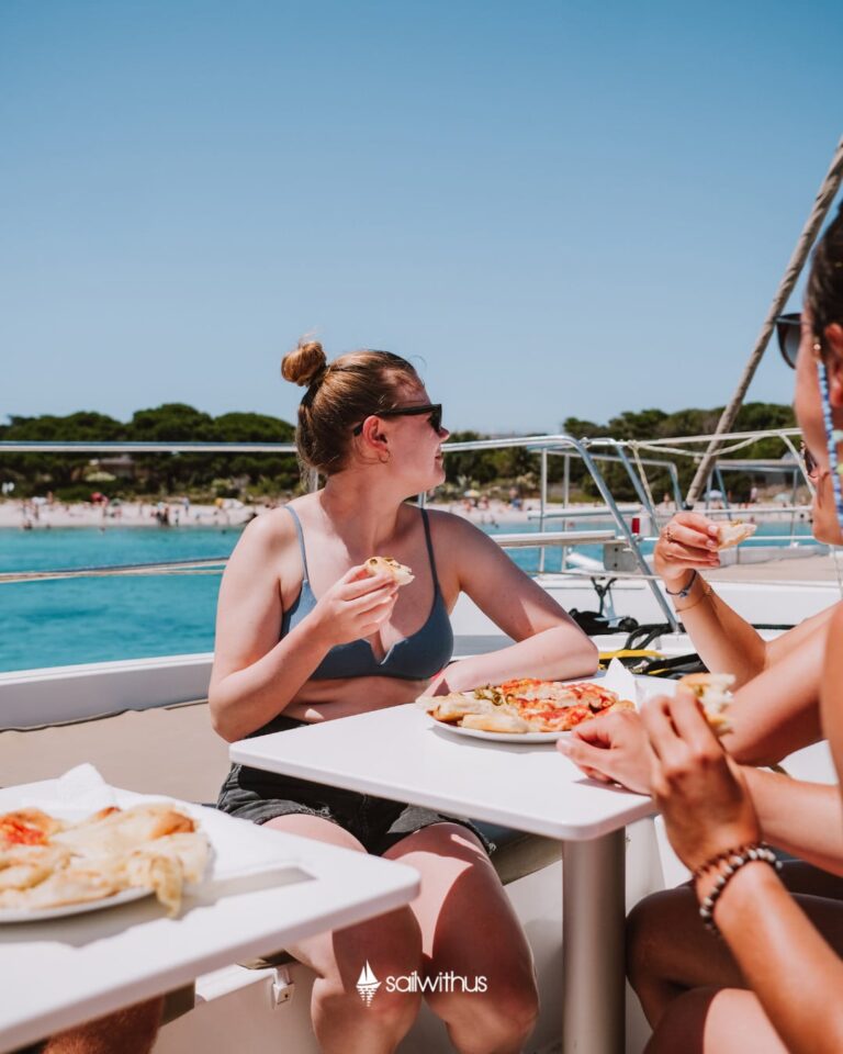 Eine Frau in Badeanzug und Shorts sitzt an einem Tisch im Freien auf einem Boot und genießt mit ihren Begleitern während ihres Segelurlaubs Pizza. Sie sind umgeben von klarem blauen Wasser und einem Sandstrand mit Bäumen im Hintergrund. Der Himmel ist klar und sonnig, was für einen perfekten Tag zum Mitsegeln sorgt.