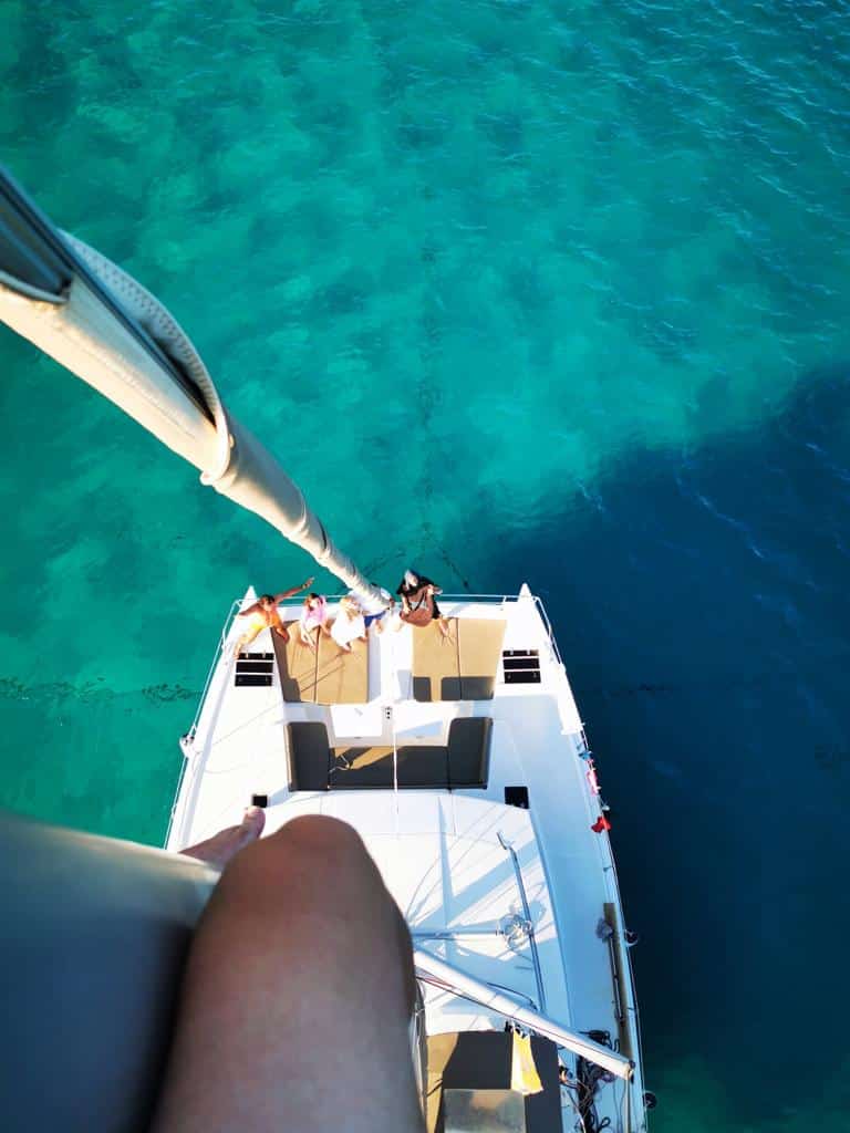Luftaufnahme von Menschen, die sich während ihrer Segelreise auf dem Deck eines weißen Bootes entspannen, das in klarem, türkisfarbenem Wasser vor Anker liegt. Im Vordergrund sind der hohe Mast des Bootes und einige Takelagen zu sehen. Das Bein einer Person ist teilweise im Bild, aus einer höheren Perspektive.