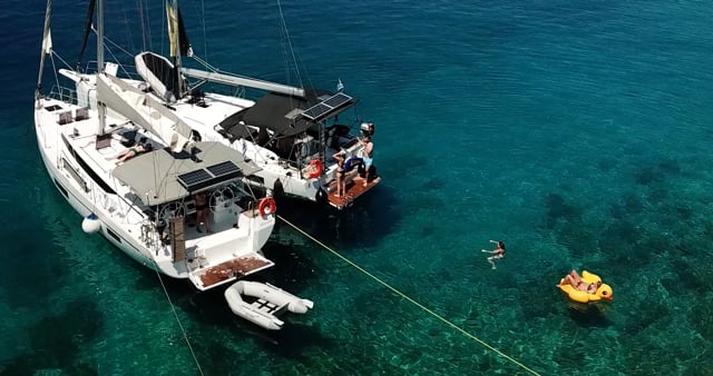 Two sailboats, including a sleek catamaran, lie anchored side by side in clear blue water. People are swimming around the boats, with one person floating on a yellow-orange inflatable raft. The scene captures a sunny, relaxing sailing holiday on the water.
